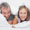 Happy middle aged couple under the duvet smiling at camera in bedroom at home