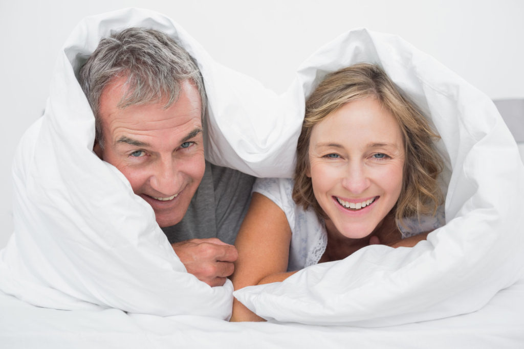 Happy middle aged couple under the duvet smiling at camera in bedroom at home
